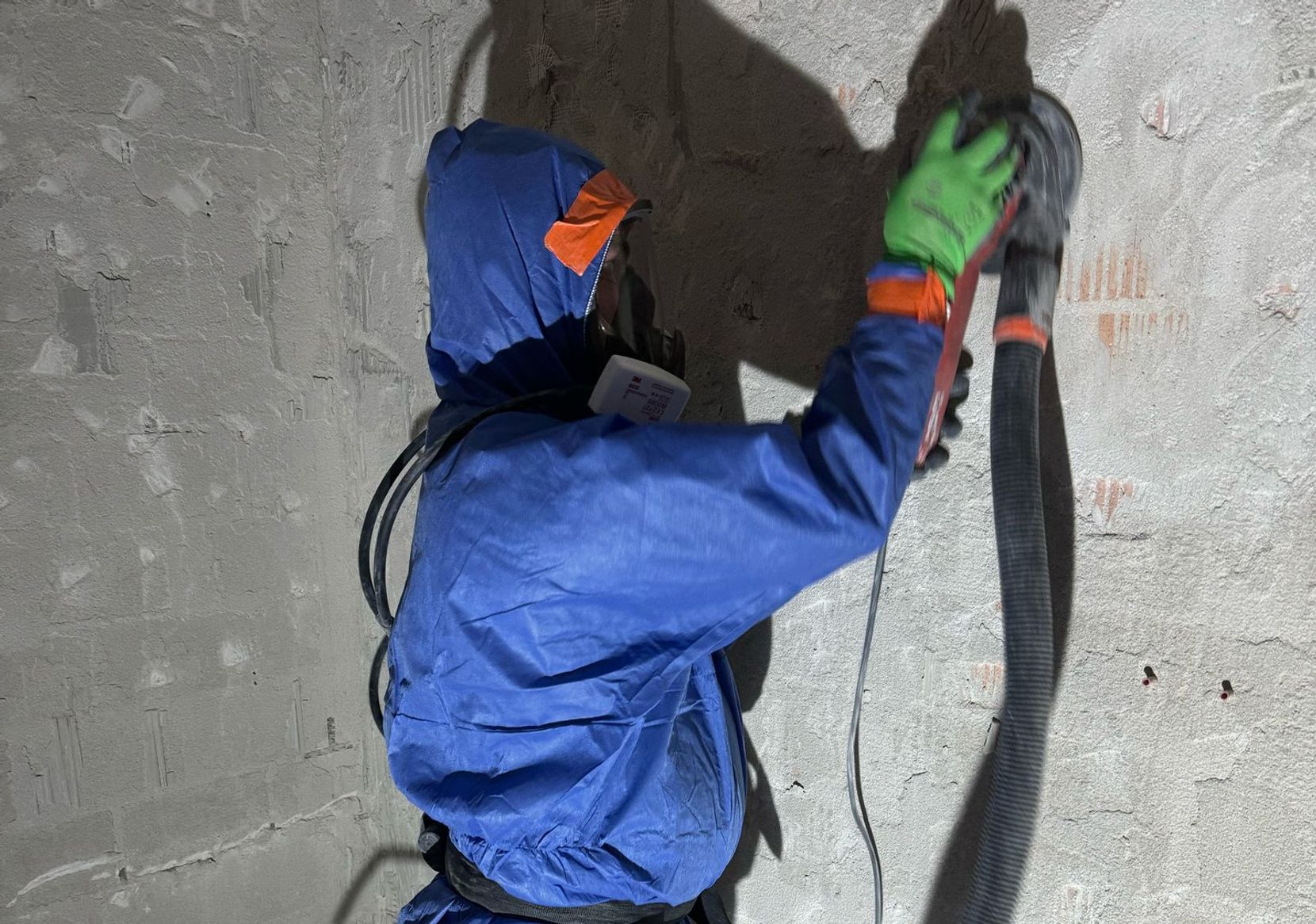 Ausgerüstet mit einem Ganzkörperanzug, Stiefeln und einer Maske, durch die sie mit frischer Luft versorgt werden, schleifen die Arbeiter das Asbest von den Wänden. 