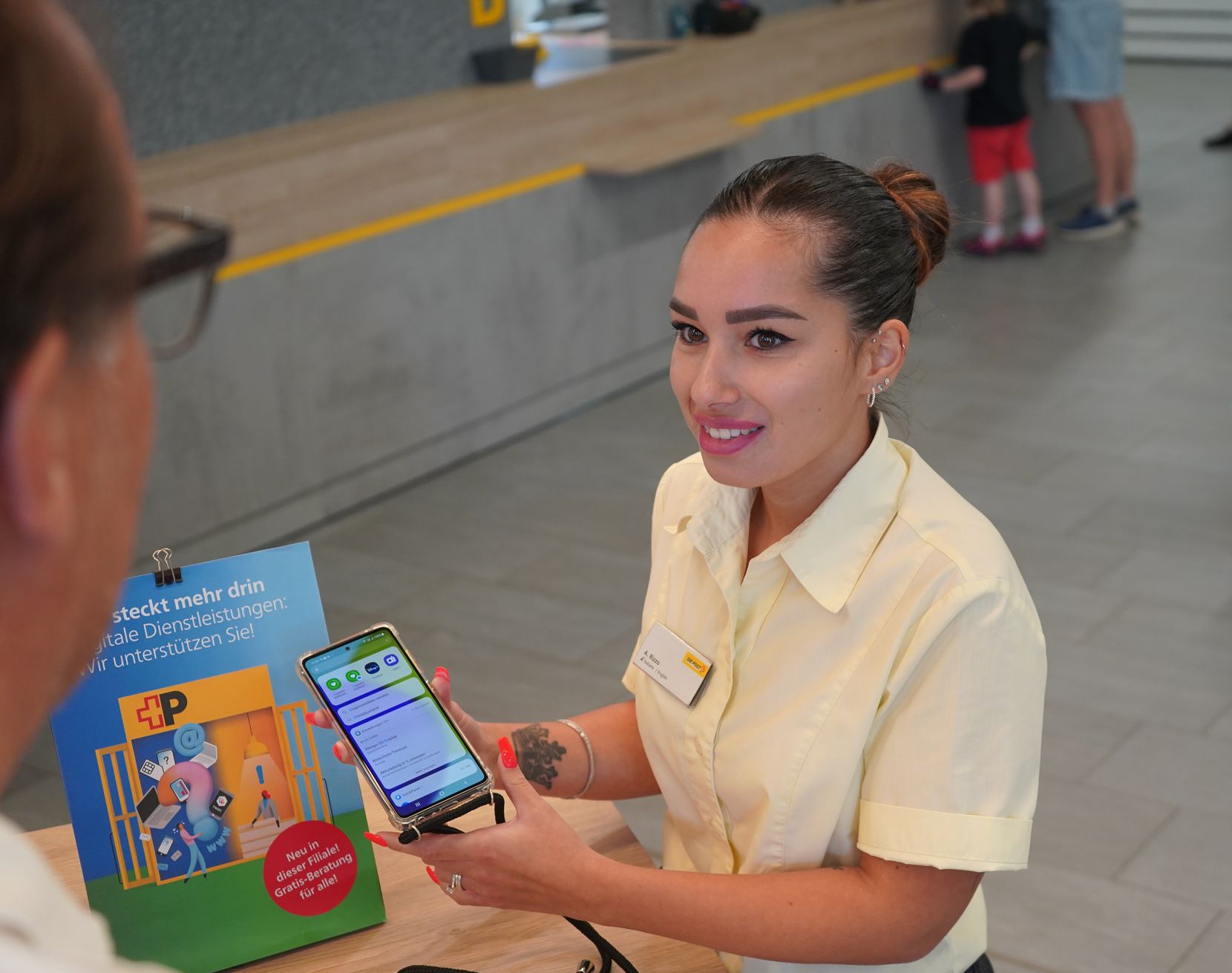 Eine junge Postangestellte hält ein Smartphone in der Hand und erklärt einem Kunden etwas.