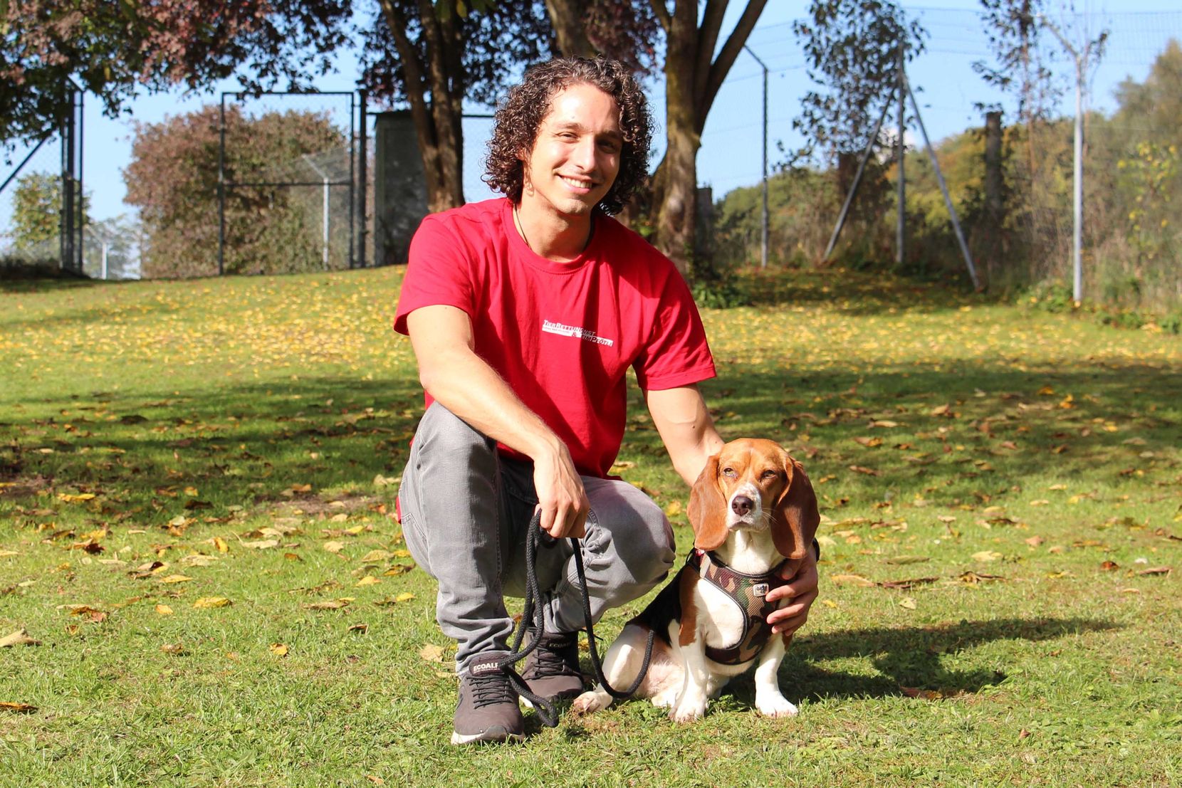 Ein junger Mann kauert neben einem Hund auf einer Wiese und lächelt in die Kamera.