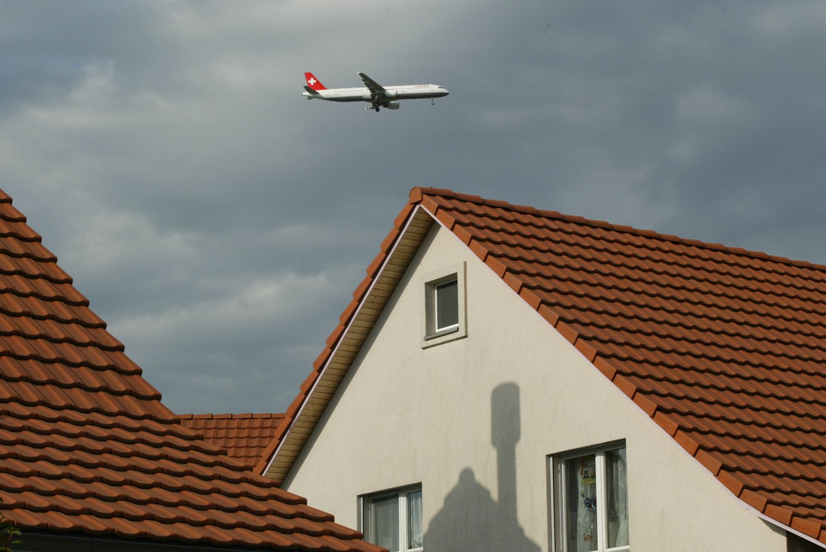 Häuser mit Giebeldach, darüber ein Passagierflugzeug im Landeanflug. 