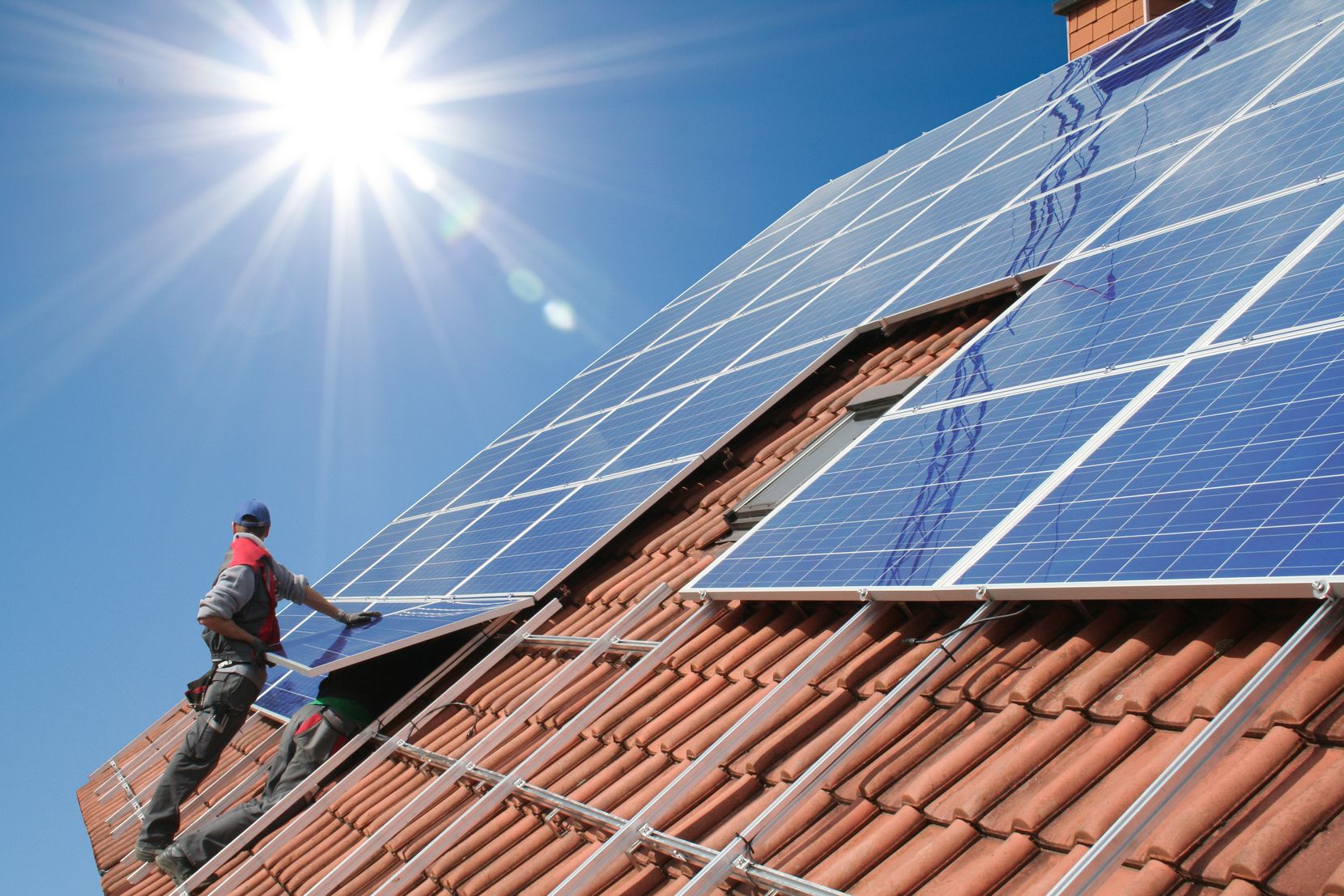 Auf dem Bild ist ein Mann auf einem Dach an einer Photovoltaikanlage werkelnd zu sehen.