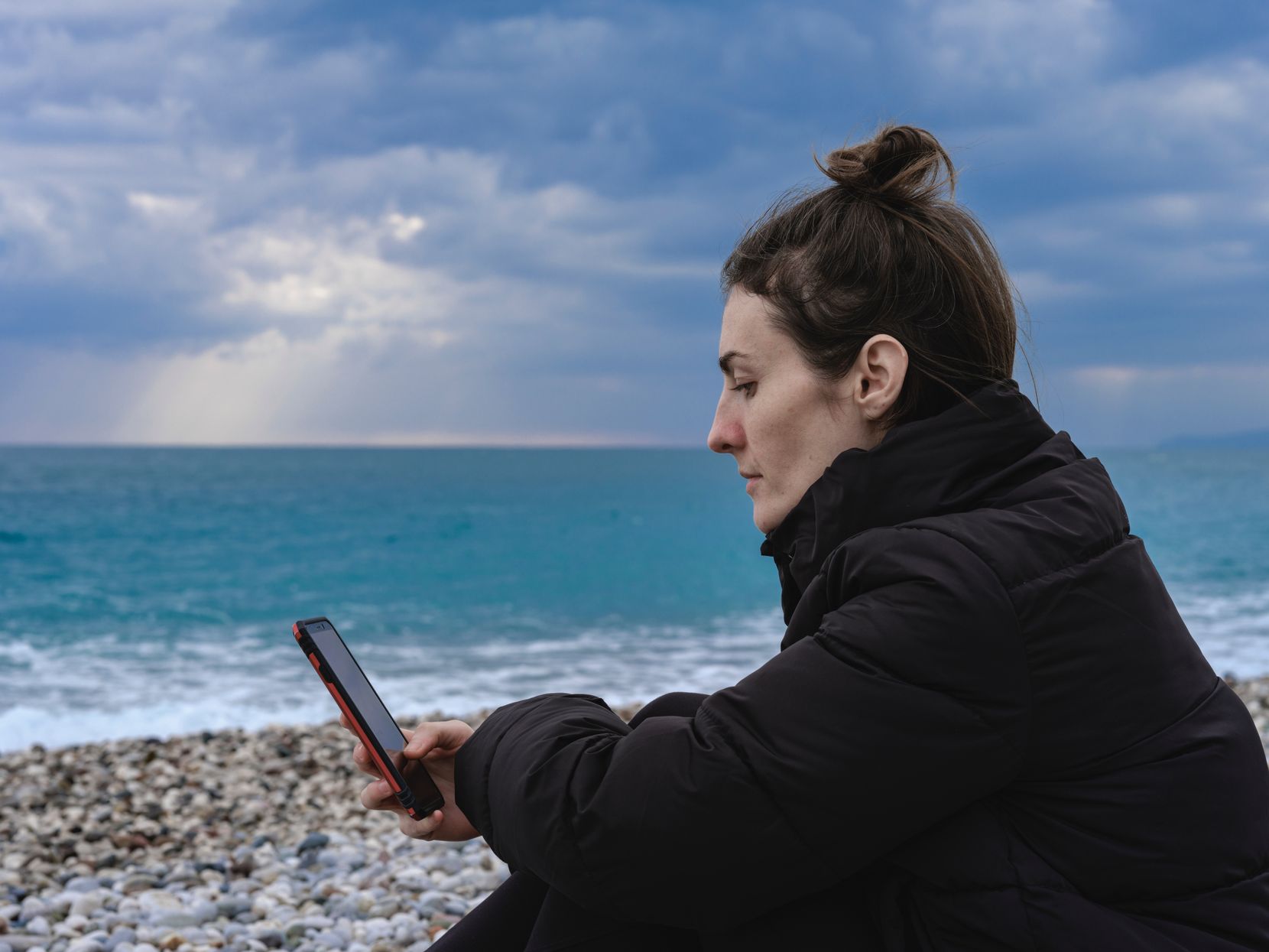 Eine Frau sitz angezogen am Meer und blickt traurig auf ihr Handy.