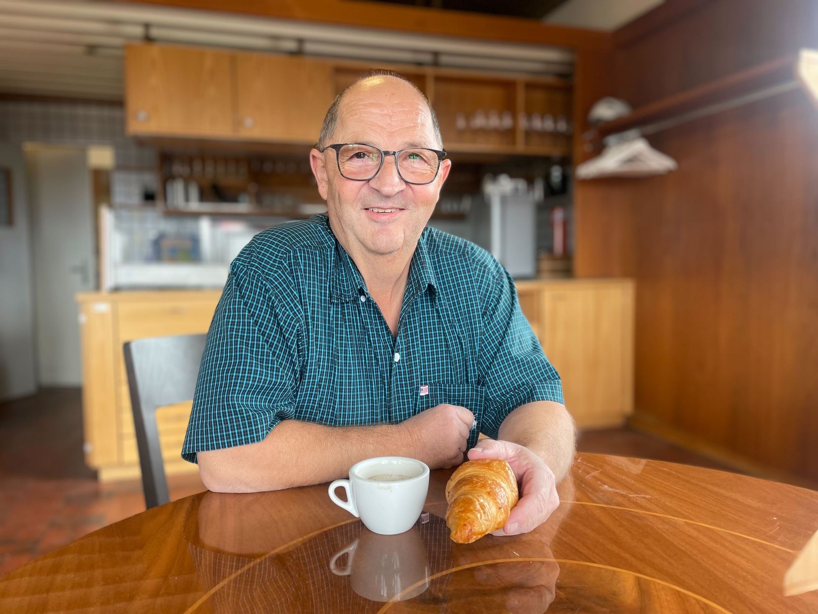 Mann mit Glatze, kariertem Hemd und Brille sitzt mit Kaffee und Gipfeli an einem Tisch.