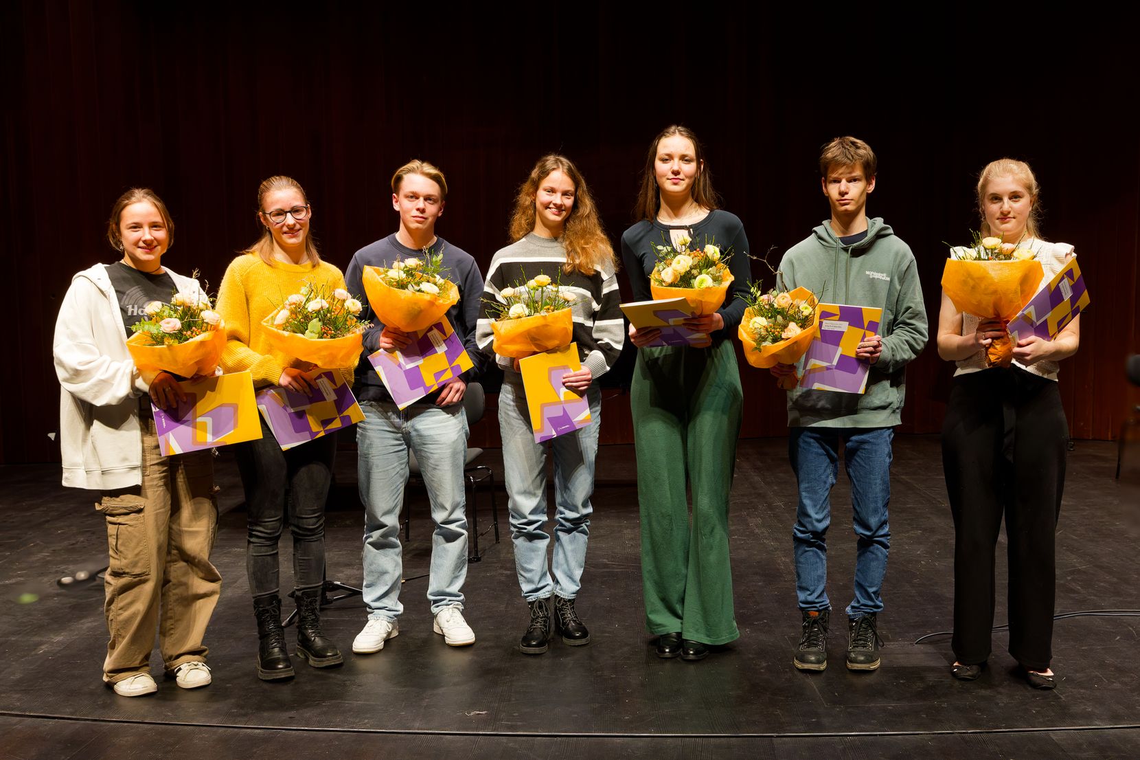 Sieben Personen stehen mit einem Blumenstrauss und einem Zeugnis auf einer Bühne im Scheinwerferlicht.