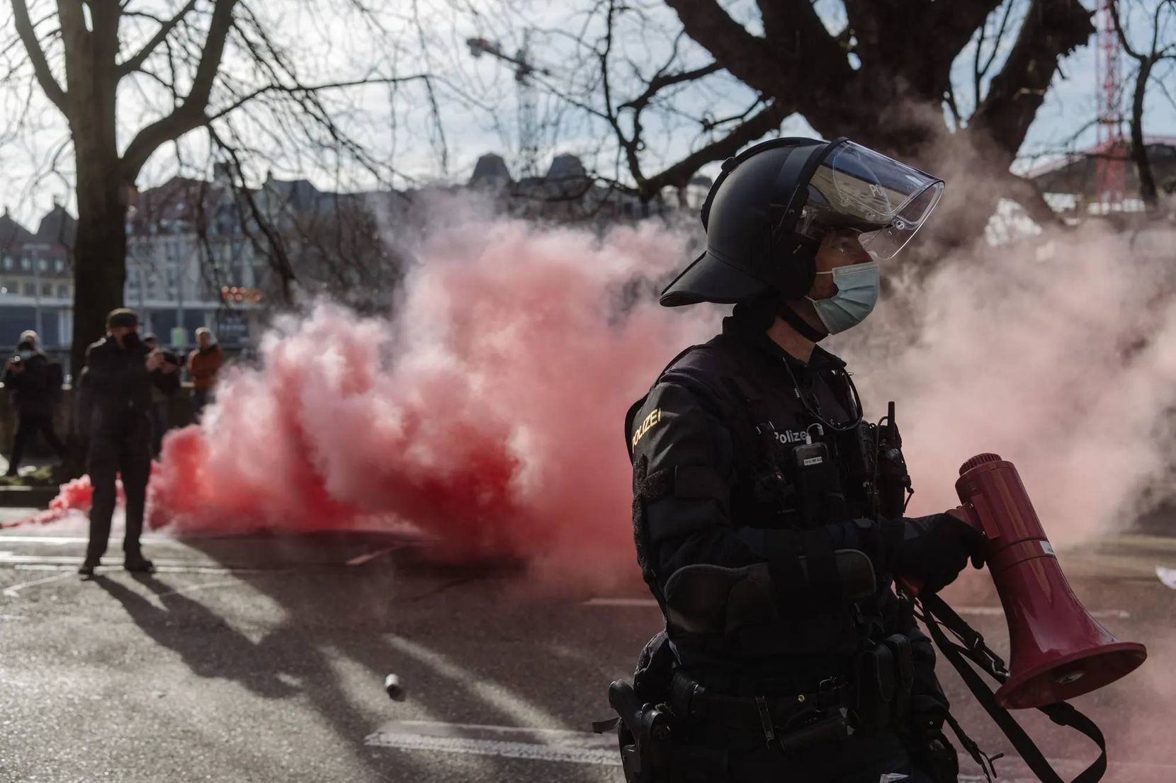 Man sieht einen Polizisten mit Megafon in ganzer Montur an einer Demonstration. Hinter ihm sieht man pinken Rauch.