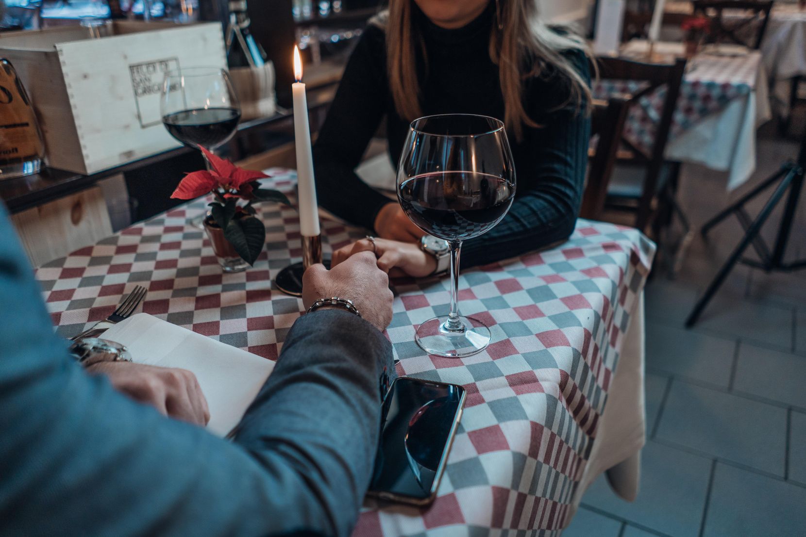 Paar, das an einem Tisch sitzt und sich an den Händen hält, jede Person ein Glas Rotwein neben sich, die Köpfe sind nicht zu sehen.