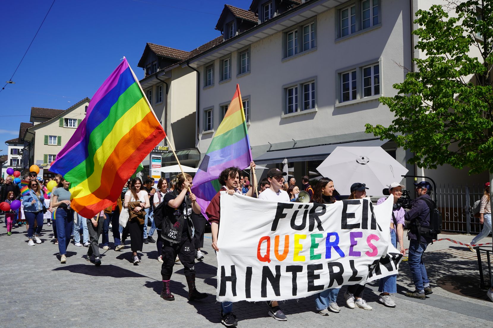 Man sieht Personen an einer Demonstration. Sie tragen ein Transparent: Für ein queeres Hinterland. Ebenfalls sichtbar sind zwei Regenbogenfahnen.
