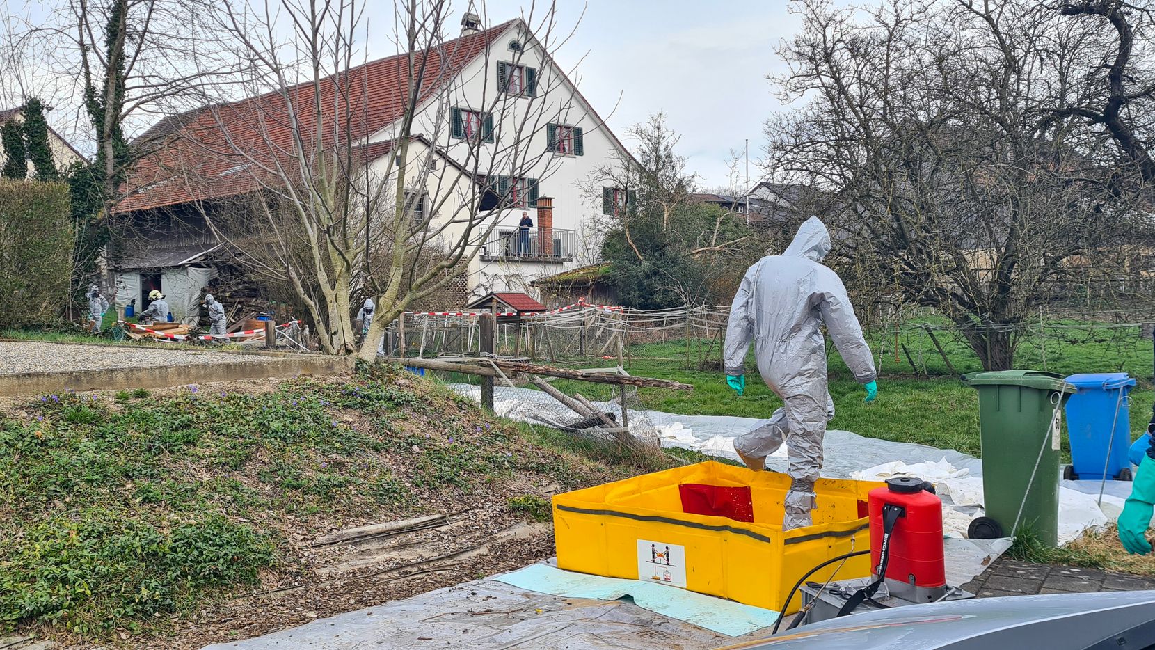 Man sieht eine Person in einem Schutzanzug bei einer Wiese. 
