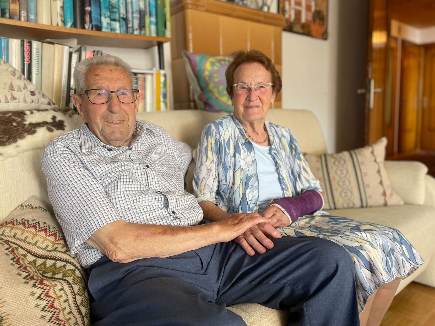 Älteres Ehepaar sitzt auf einem Sofa und schaut in die Kamera, die Frau hat einen Arm im Gips.