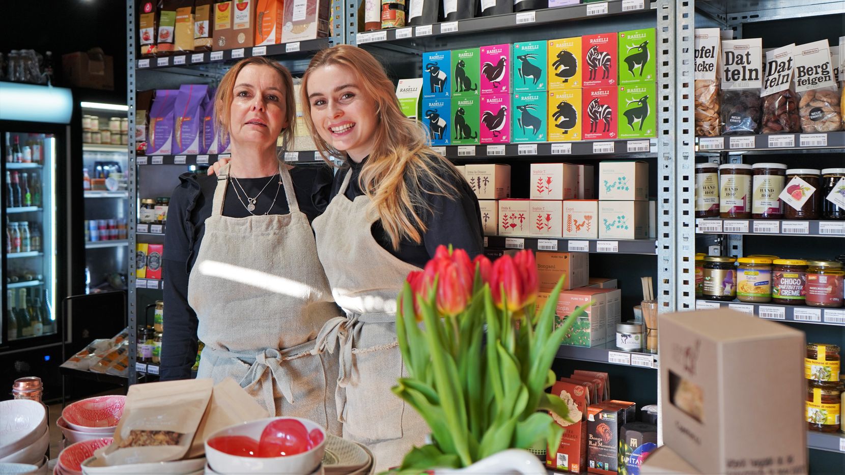 Zwei Frauen mit brauner Schürze stehen im Laden vor Produkten