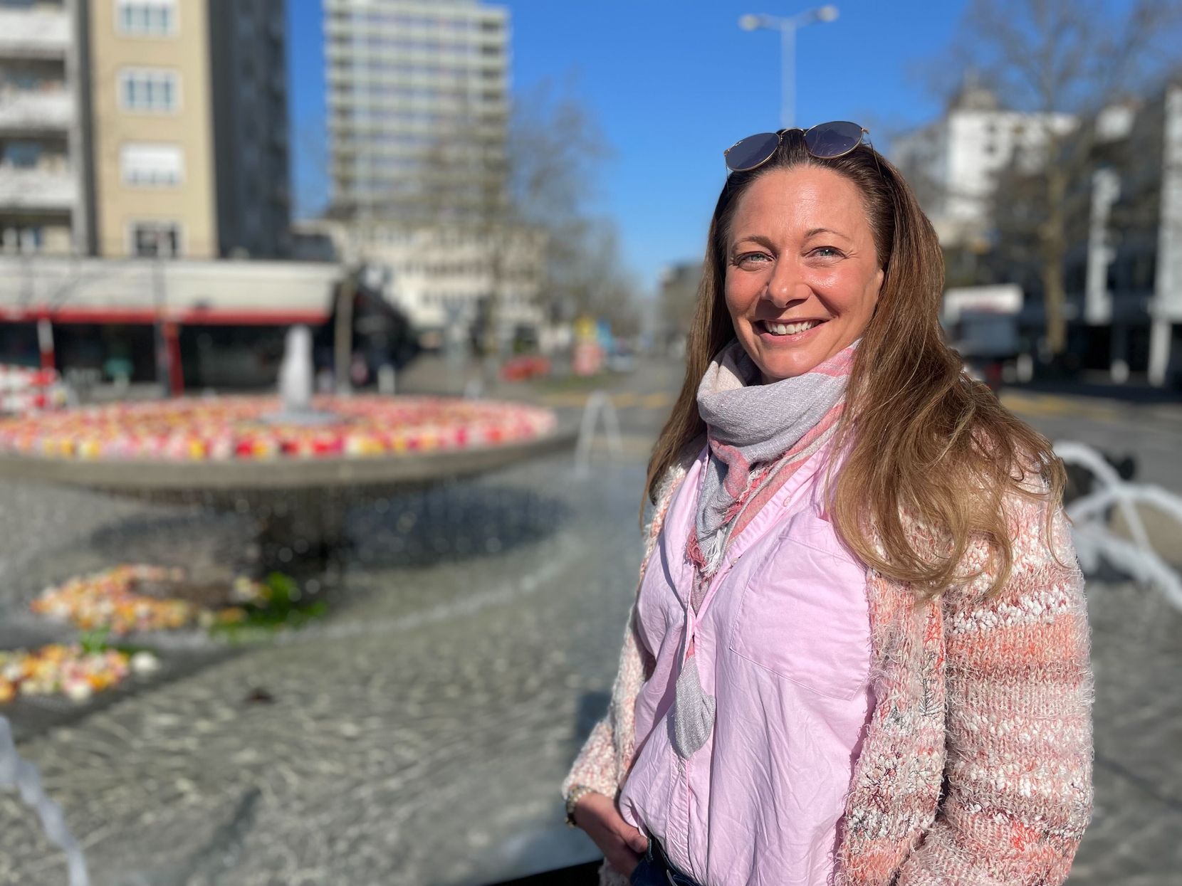 Frau mit langen Haaren, Strickjacke und pinker Bluse steht vor einem Brunnen, in dem es Rosenblätter hat.