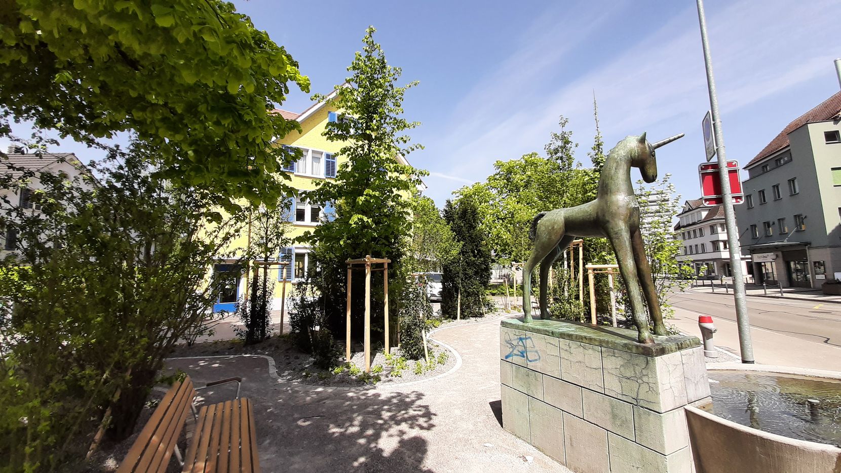 Mehrere Bäume mit Brunnen und Sträuchern auf einem kleinen Platz in einer Stadt.