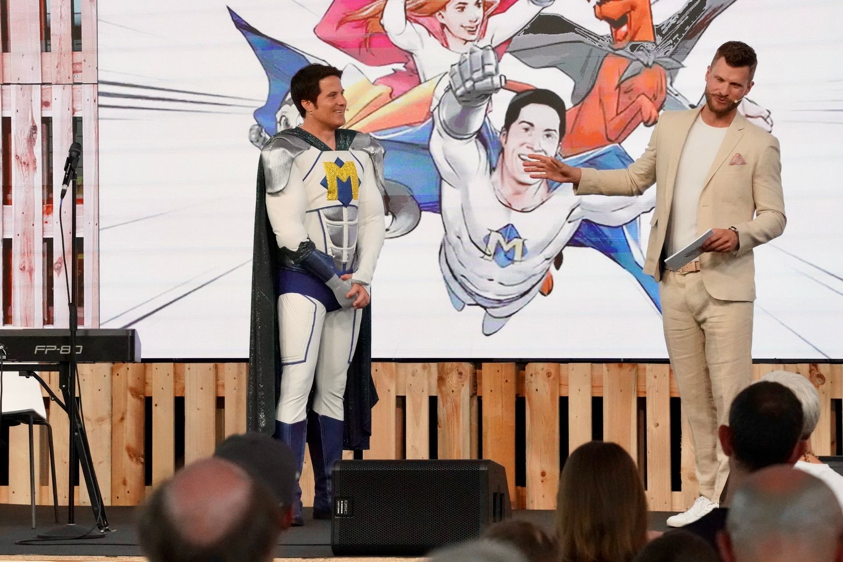 Zwei Männer, einer davon als Superheld verkleidet, stehen auf einer Bühne.