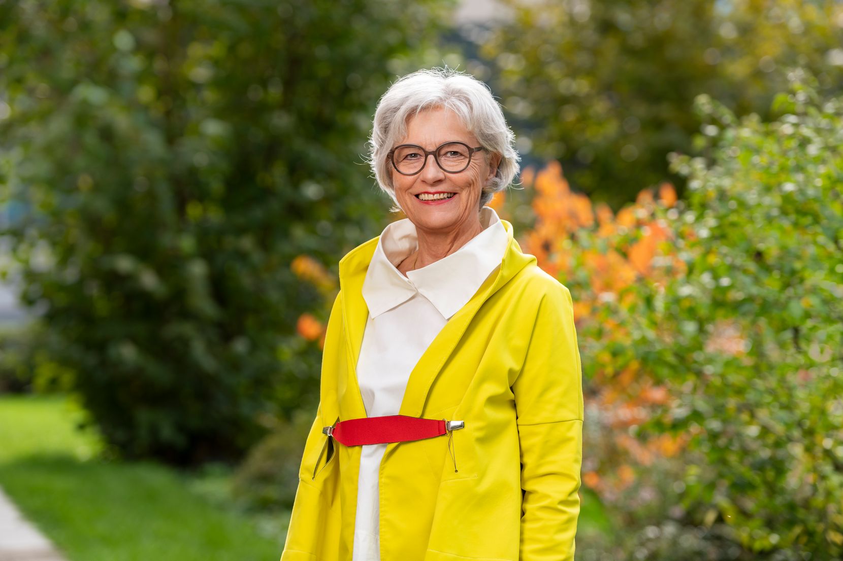 Eine ältere Frau mit kurzen weissen Haaren steht in gelber Jacke vor einer Hecke.