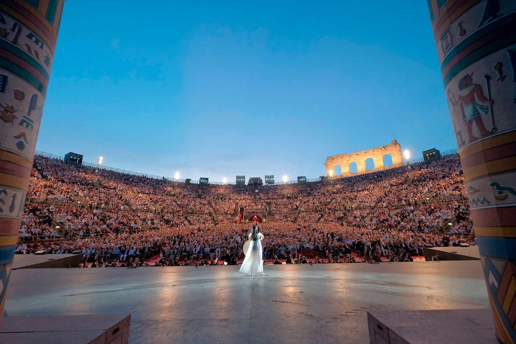 Eine Opernsängerin steht auf der Bühne in der Arena di Verona und singt zu den vollen Zuschauerrängen.