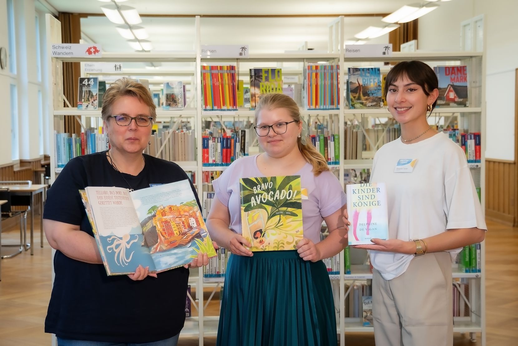 Drei Frauen stehen vor einem Bücherregal, jede hält ein Buch in den Händen. 