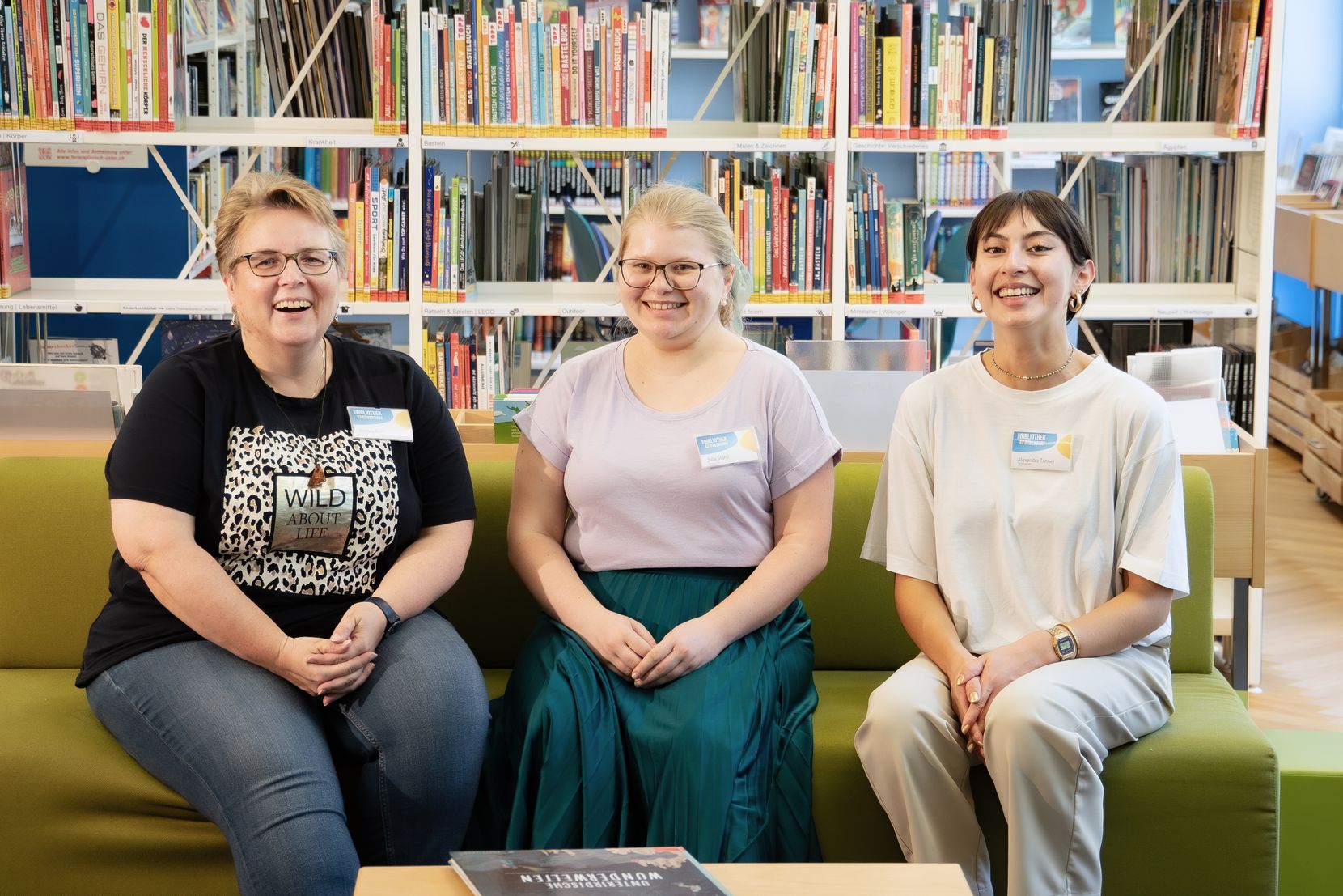 Drei Frauen sitzen auf einem Sofa in einer Bibliothek und lächeln in die Kamera. 