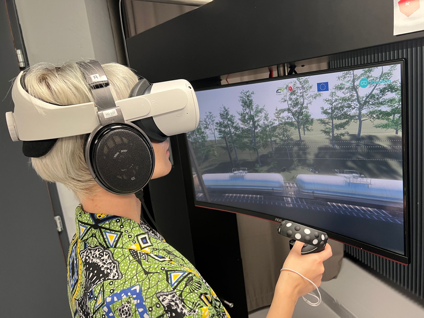 Eine Frau trägt eine VR-Brille und Kopfhörer und spielt mit einem Controller vor einem grossen Bildschirm eine Bahnlärmsimulation.