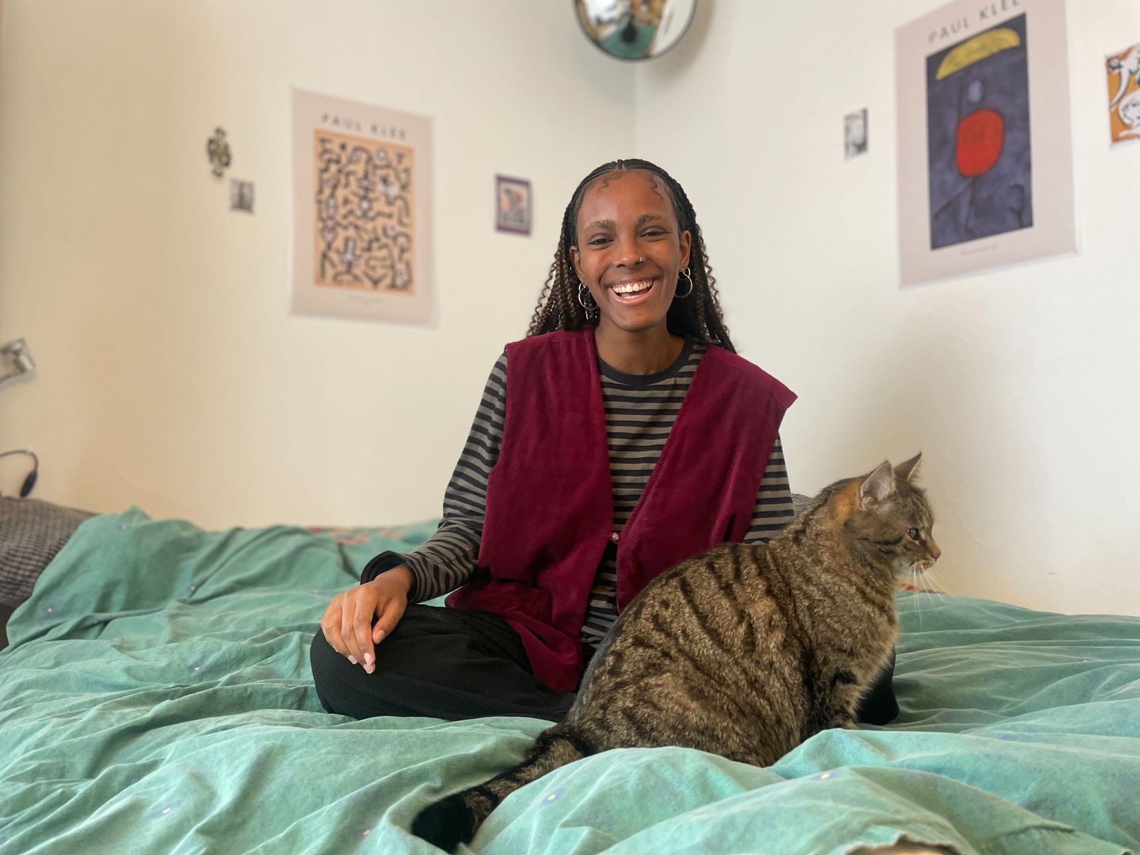 Eine junge Frau mit Zöpfchen sitzt auf ihrem Bett, vor sich eine getigerte Katze.