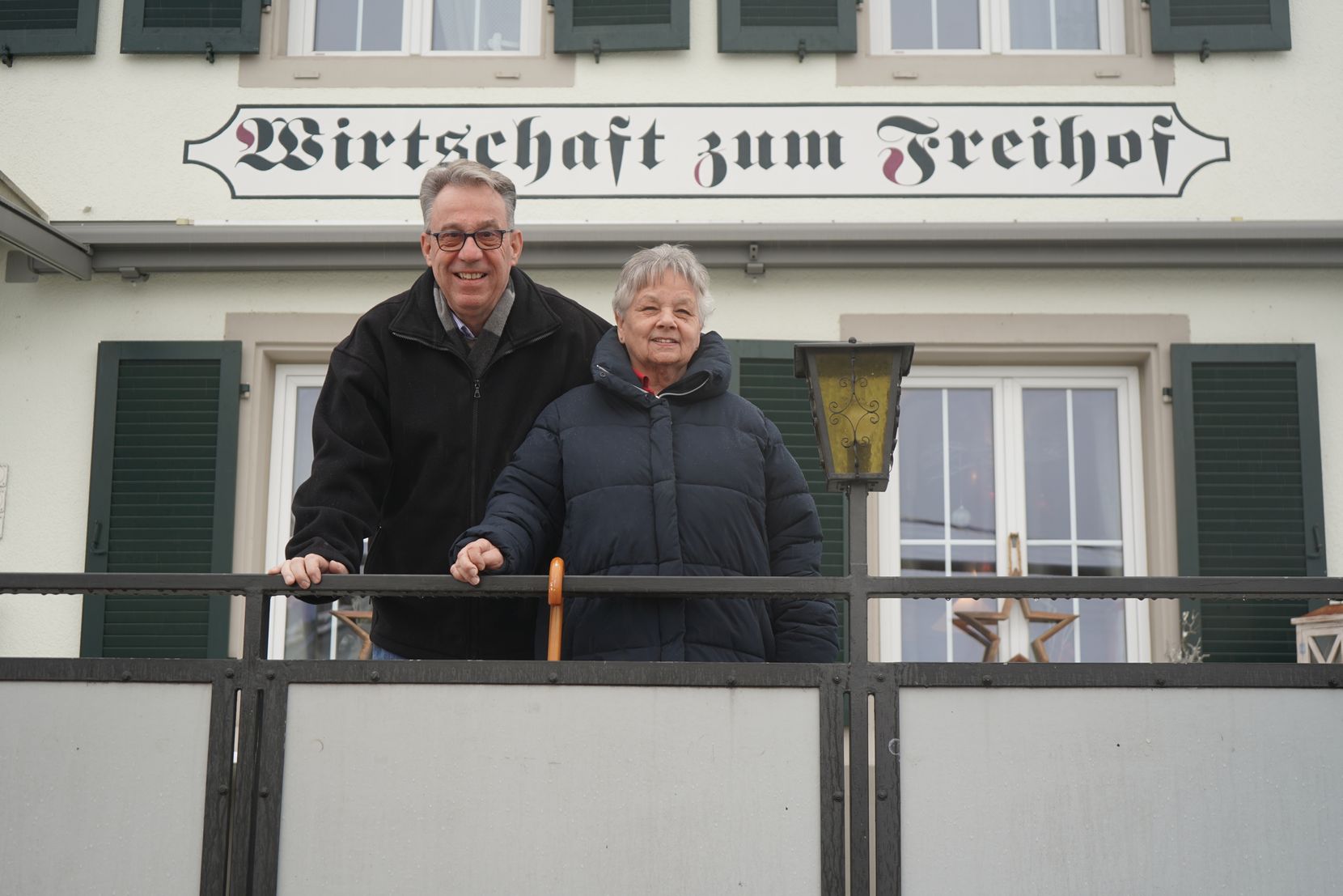 Ein Mann und eine Frau stehen auf der Terrasse eines Restaurants. Im Hintergrund das Schild «Wirtschaft zum Freihof».