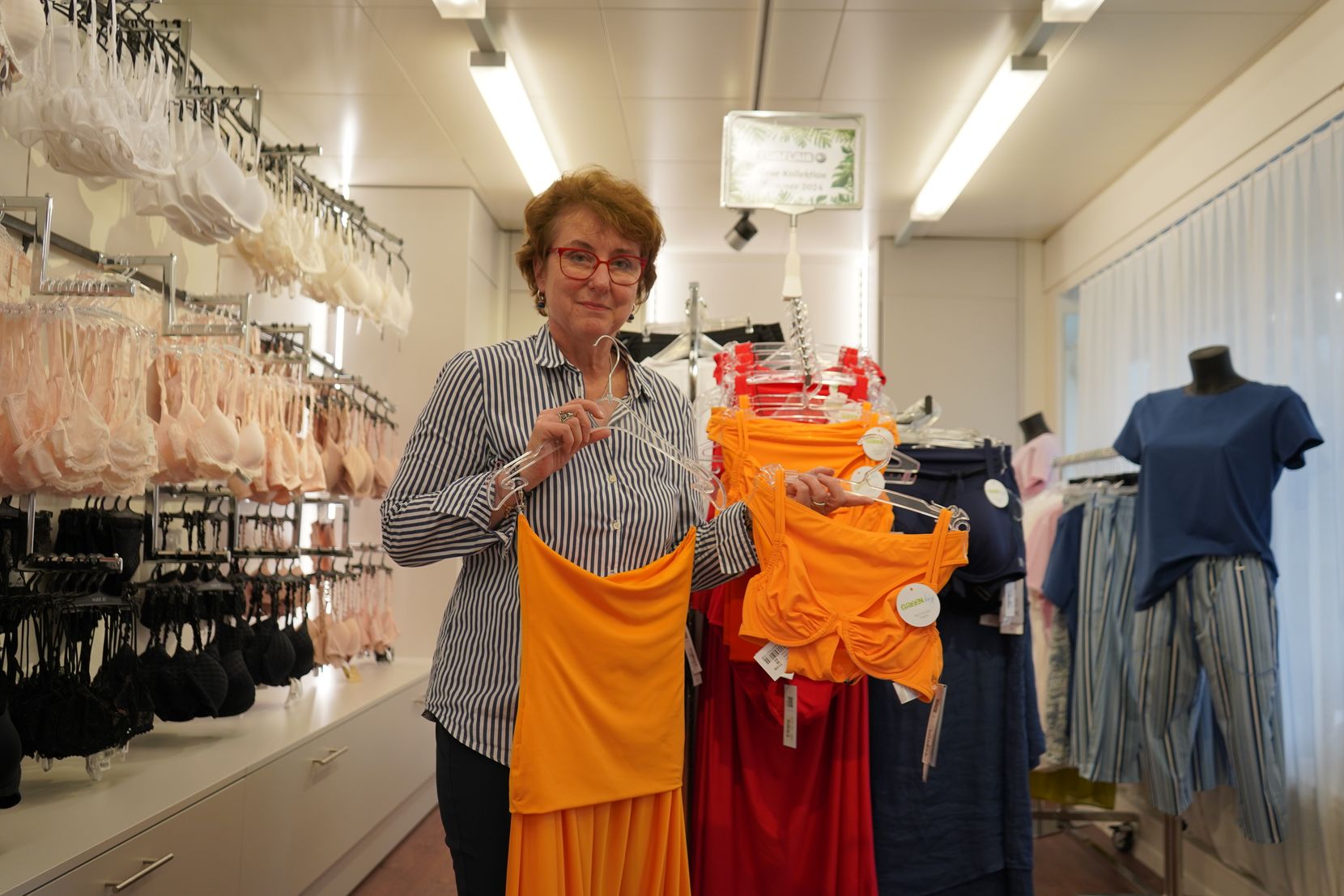 Eine Frau in einer blauen Bluse hält in ihrem Geschäft für Dessous- und Bademode Kleidungsstücke in die Kamera.
