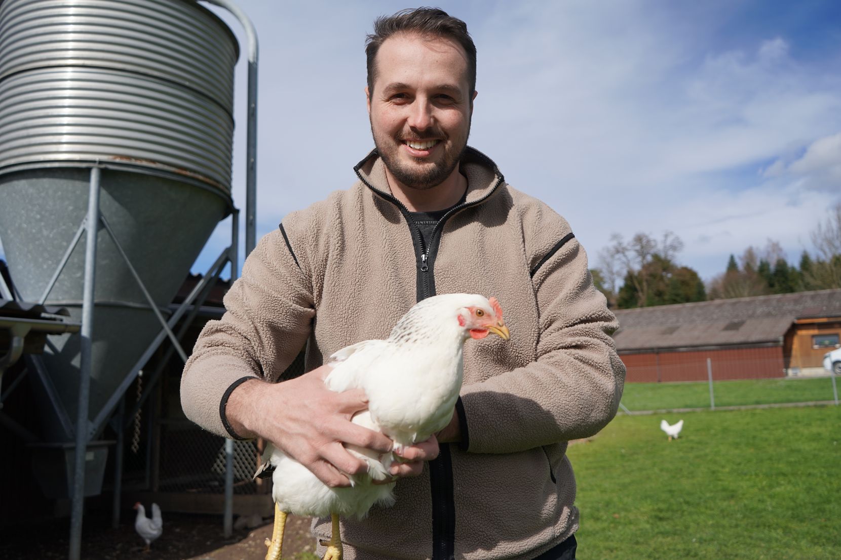 Ein Mann hält ein weisses Huhn in der Hand und lächelt in die Kamera.