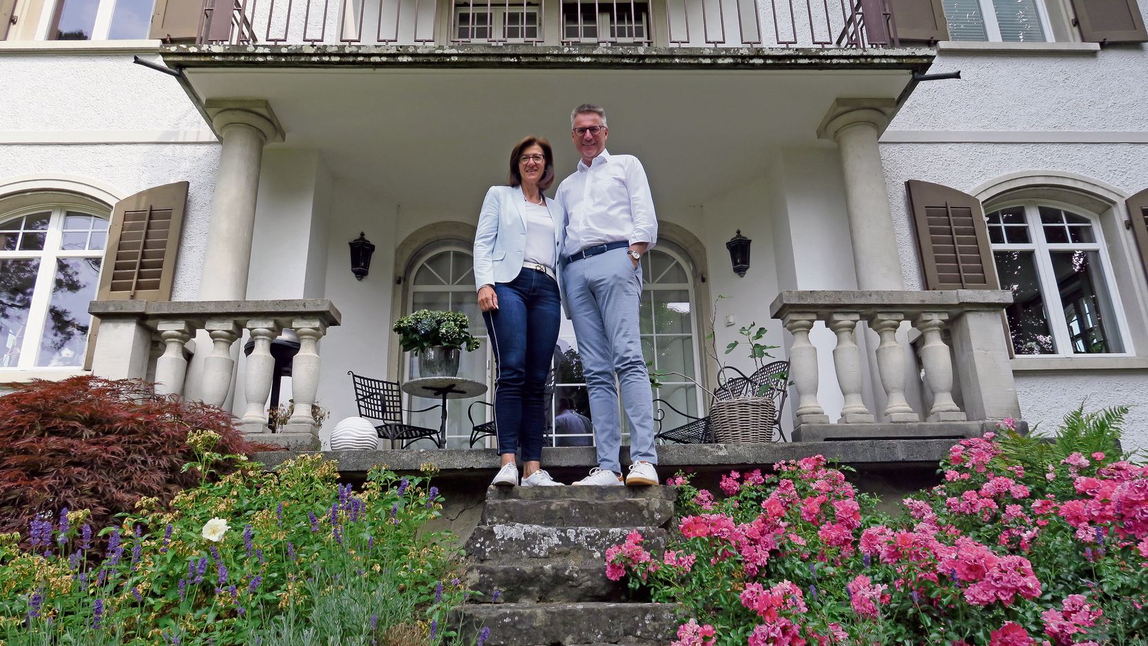 Ein Ehepaar steht auf einer Treppe, die zur Veranda des Wohnhauses führt, und lächelt in die Kamera.