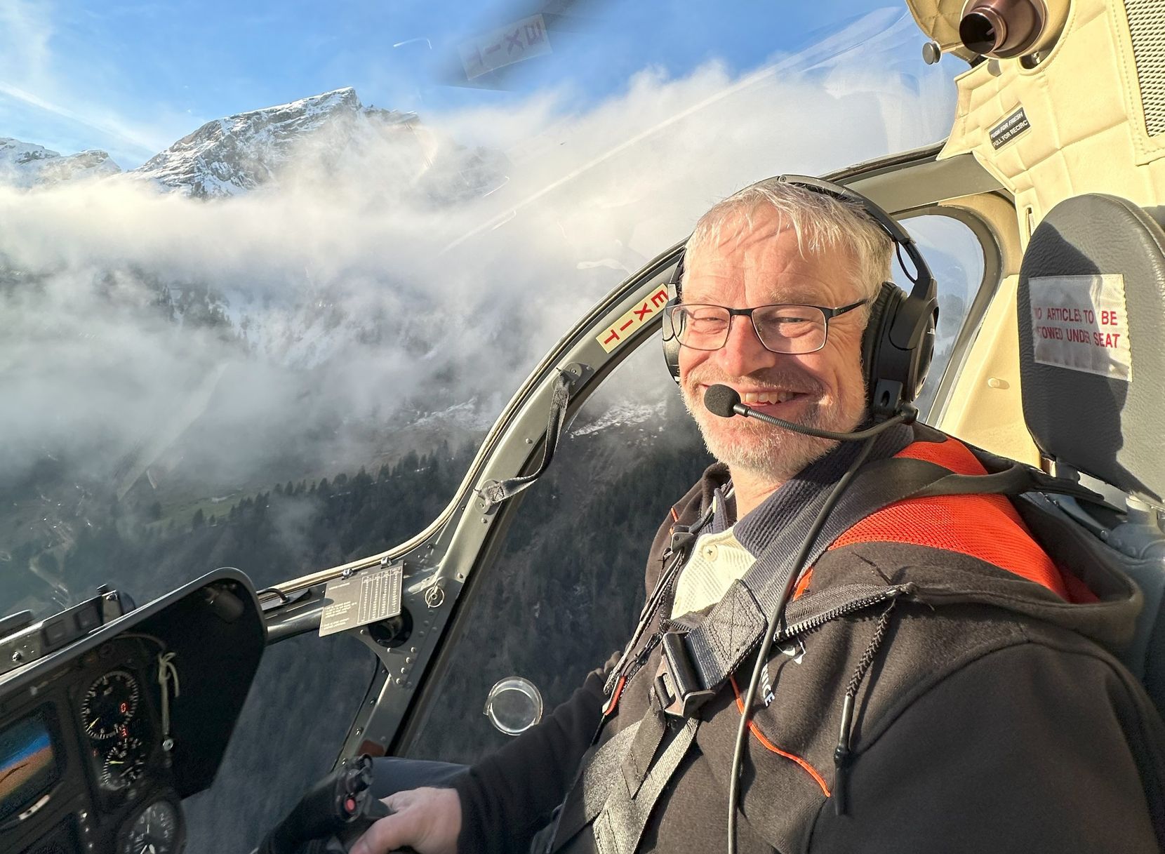 Ein Mann fliegt einen Helikopter. Im Hintergrund sieht man verschneite Berge und ein paar Wolken.