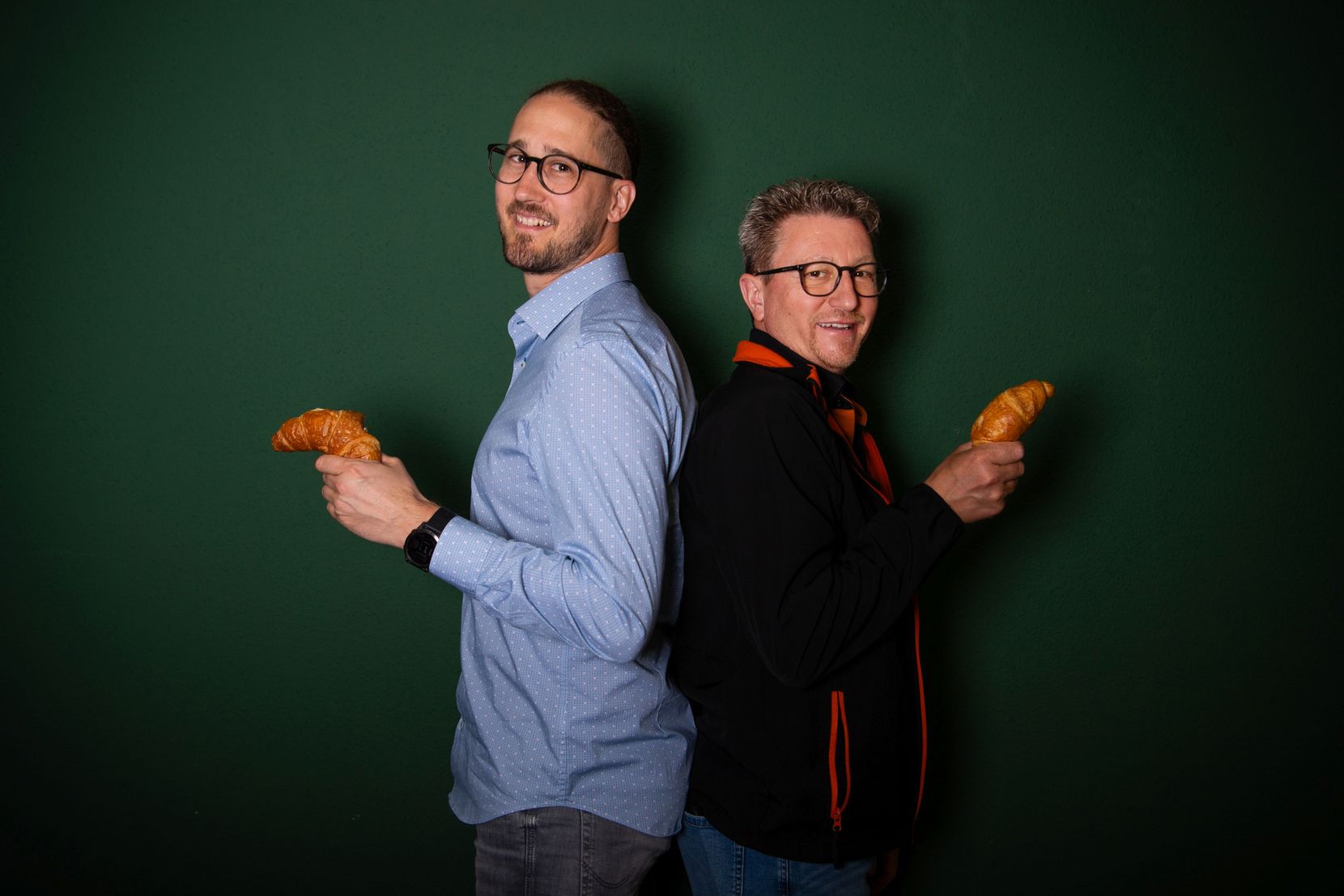 Die Bäcker Martin Mayer und René Schweizer stehen Rücken an Rücken und halten je ein Gipfeli in der Hand.
