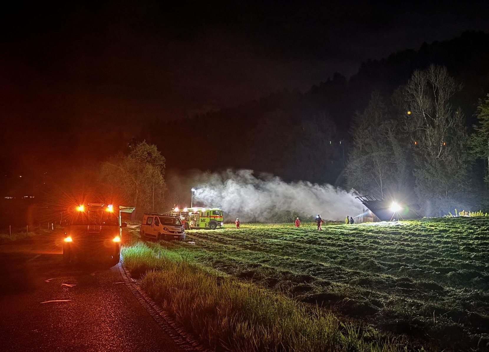 Man sieht ein Feld bei Nacht, ein Gebäude mit Rauchschwanden und Feuerwehrautos.