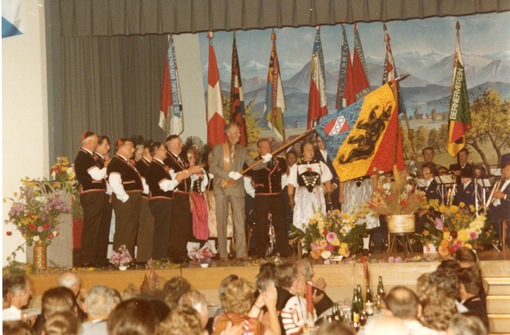 Man sieht eine Gruppe von Menschen in der Tracht auf einer Bühne stehen. Ein Mann schwenkt eine Fahne.