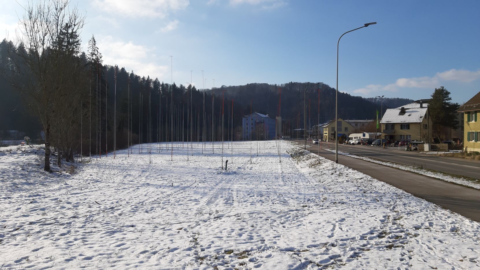 Man sieht eine schneebedeckte Wiese mit Bauvisieren darauf. Links daneben ist ein kleiner Wald, rechts daneben die Kantonsstrasse.