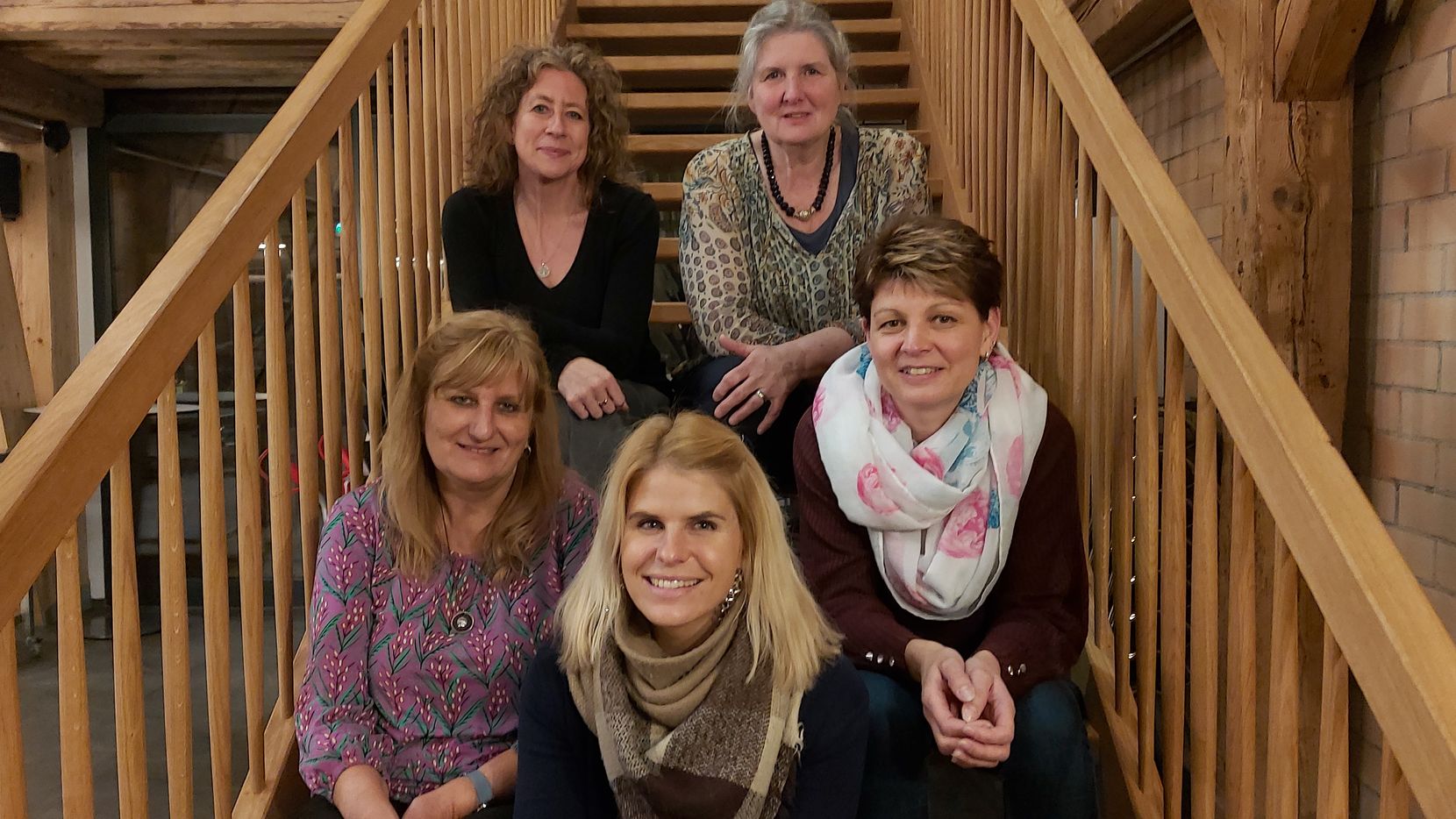 Man sieht fünf Frauen mittleren Alters, die auf einer hölzernen Treppe sitzen.