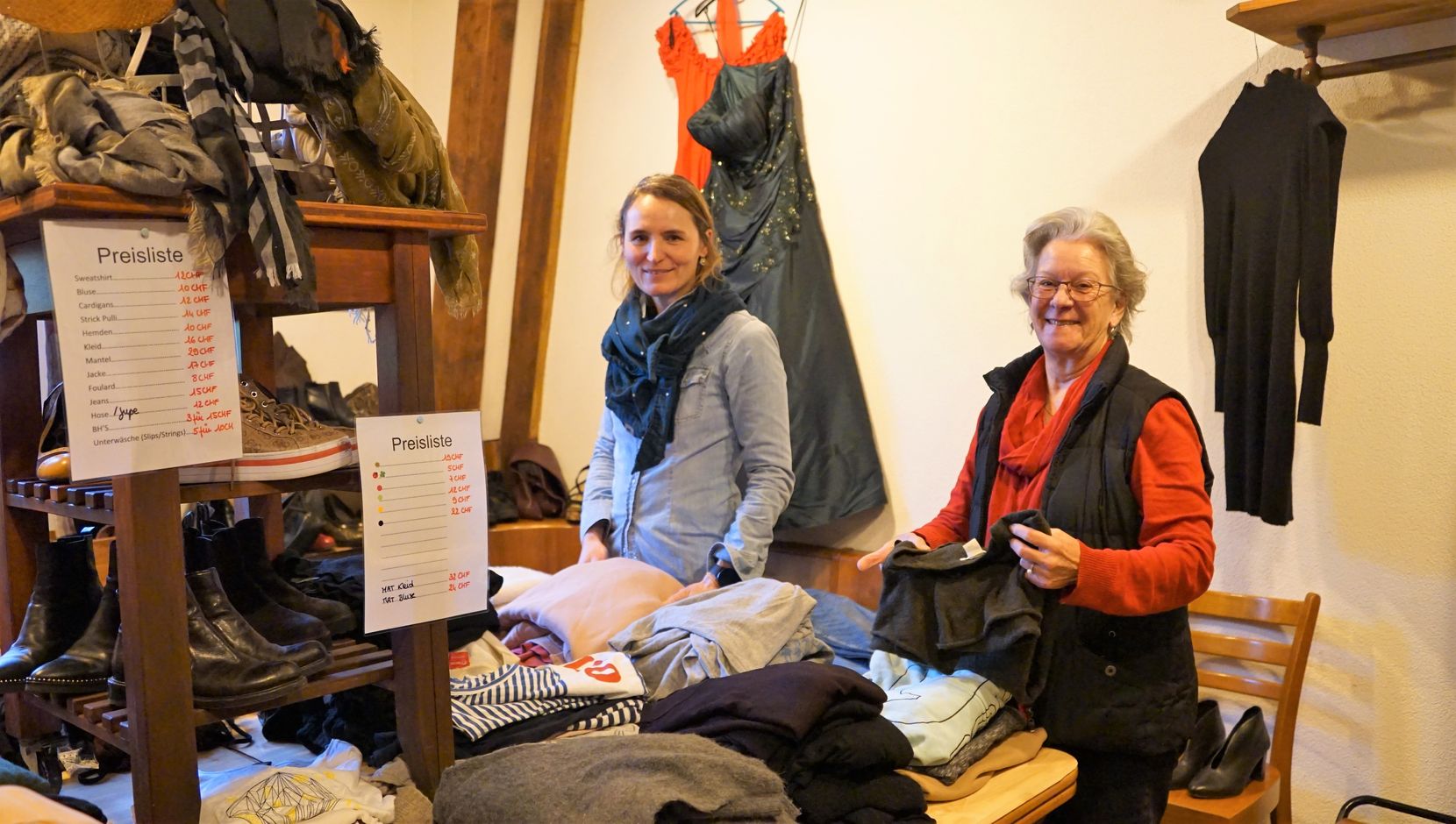 Man sieht zwei Frauen in einem improvisierten Second-Hand-Shop. Vor ihnen liegen viele Kleider.