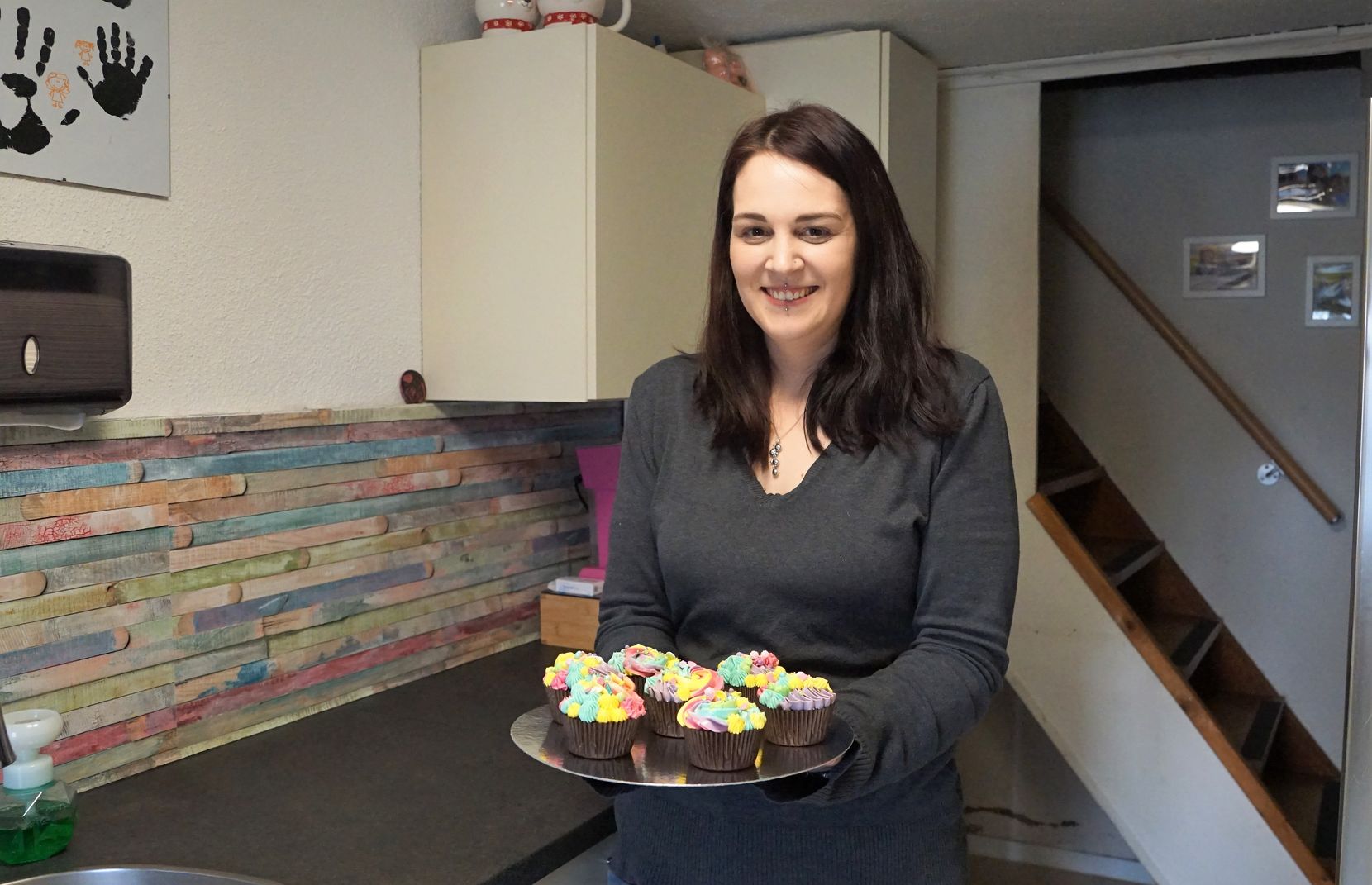 Man sieht eine Frau in einer Küche, die einen Teller mit schön dekorierten Cupcakes trägt.