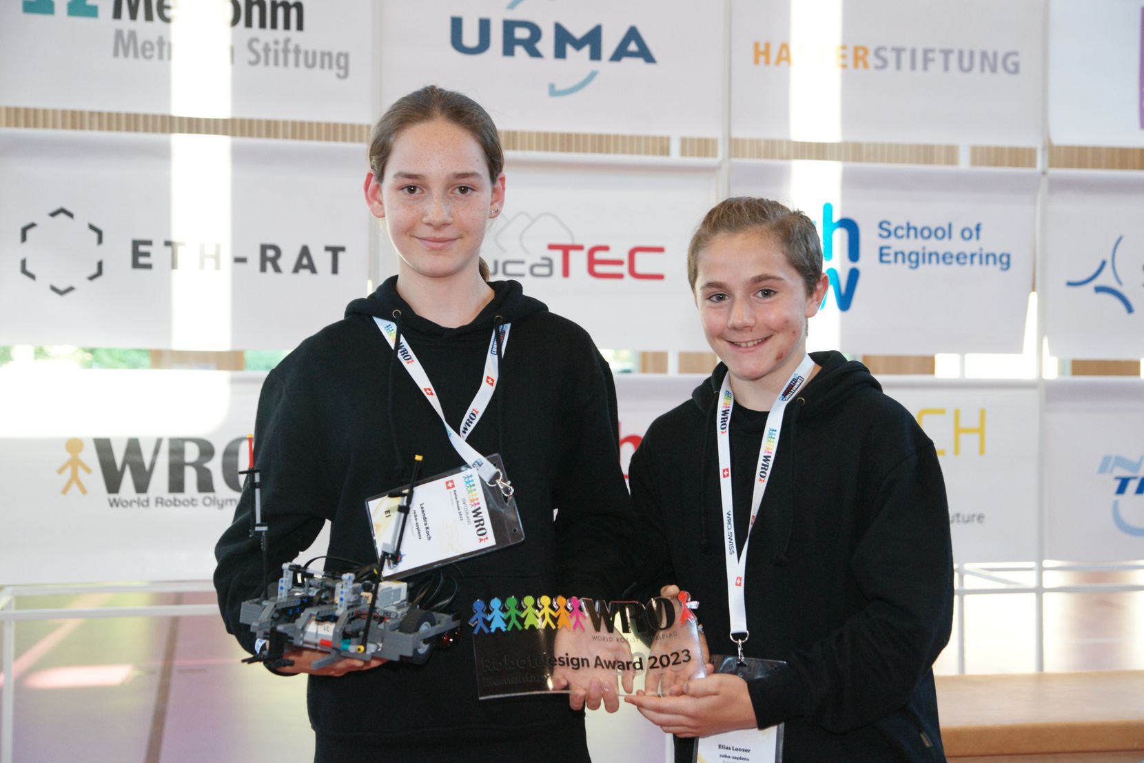 Ein Mädchen hält einen Roboter in der Hand. Neben ihr steht ein Junge mit einem Award.