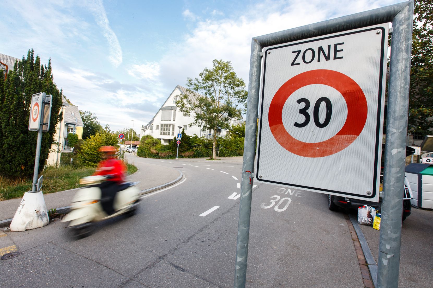 Strasse mit Tempo-30-Schild und einem vorbeifahrenden Vespa-Fahrer. 