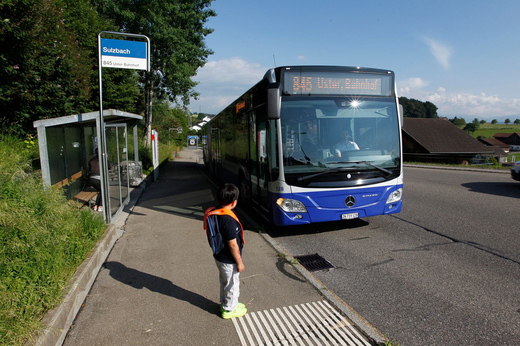 Man sieht die Bushaltestelle Sulzbach in Uster, wo ein Kind auf den anfahrenden Bus wartet.