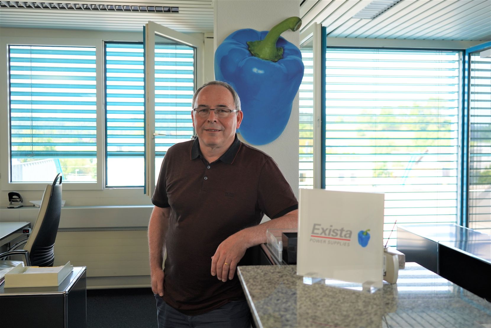 Ein Mann lehnt mit dem linken Arm an ein Büromöbel. Im Hintergrund sieht man eine blaue Peperoni an der Wand – das Logo des Fehraltorfer KMU Exista AG.