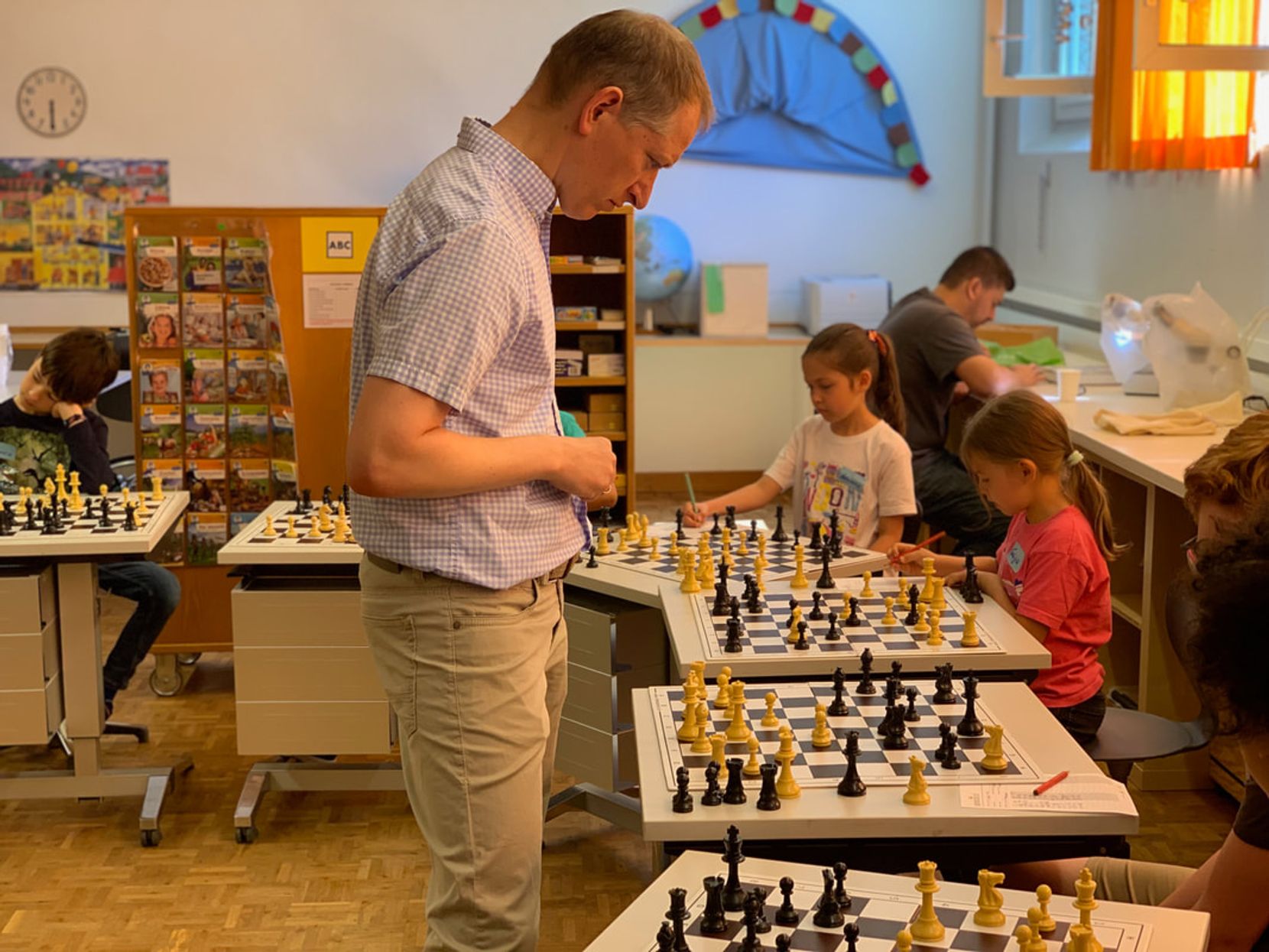 Auf dem Bild ist ein älterer Mann zu sehen, der vor an Schachbrettern sitzenden Kindern steht.