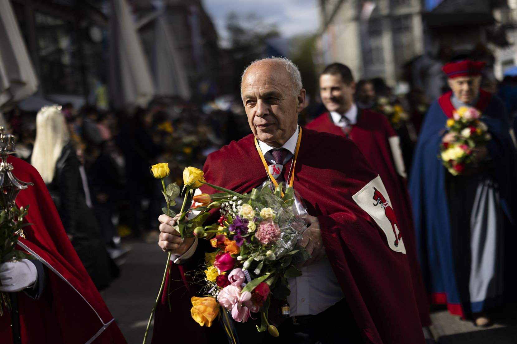 Ein alter Mann mit weissen Haaren und einem roten Umhang mit Wappen hält einen Blumenstrauss in der Hand.