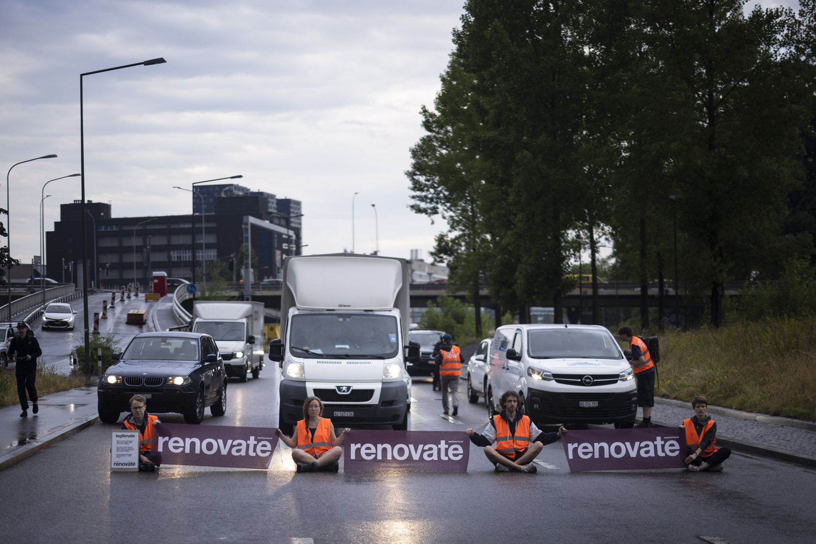 Vier Personen sitzen mit Transarenten mit der Aufschrift «Renovate» auf einer Strasse. 