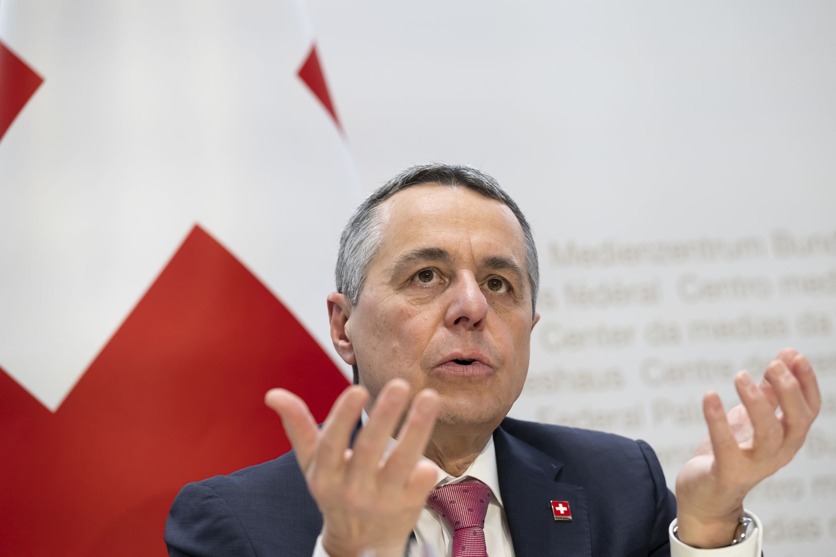 Ein Bundesrat sitzt auf einem Podium vor der Schweizer Flagge.