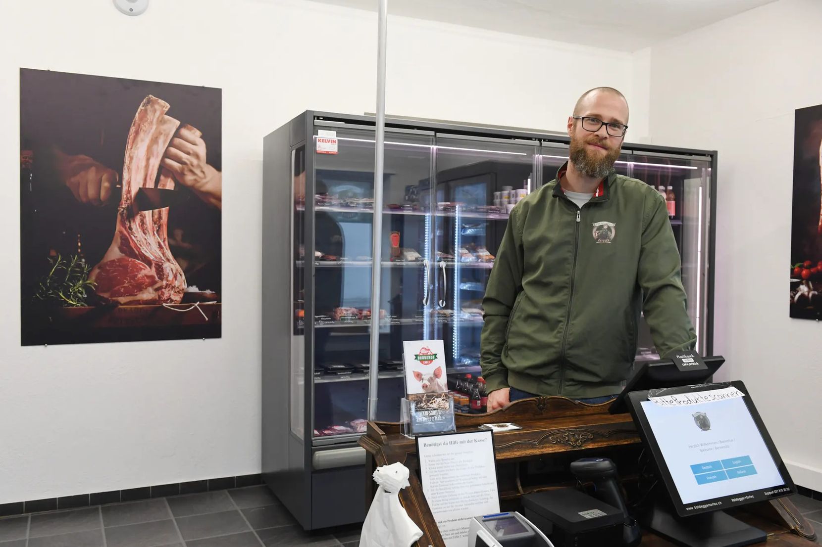 Mann mit grüner Arbeitskleidung steht hinter einer Fleischtheke.